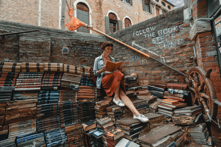 A woman is shown reading while sitting on a large stack of books.